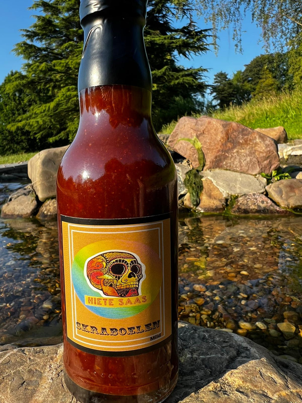 Sauce piquante Skraboelen au chipotle et piment ghost 200ml, bouteille de sauce BBQ épicée en plein air, devant un ruisseau.